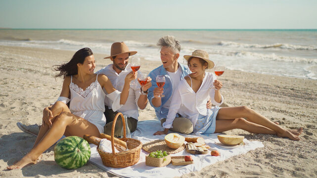 Happy group of friends enjoying beach picnic together, sharing conversation, drinking wine, celebrating weekend reunion gathering