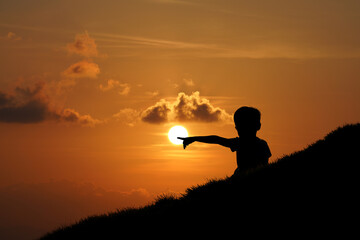 Silhouette of a boy pointing to the sun on the hill with sunset sky background.