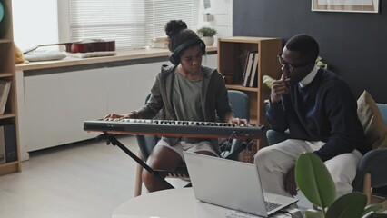 Medium long of talented girl wearing earphones, learning to play synthesizer with young Black male tutor sitting on couch in modern apartment at daytime