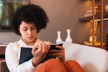 focused black woman reading book and sitting in armchair at home