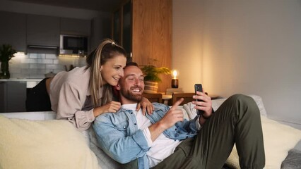 Happy caucasian couple sitting on couch taking selfies with cellular device 