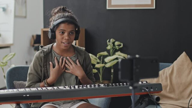 Medium of eleven-year-old Black girl sitting in armchair in modern apartment at daytime, playing synthesizer, waving hand, smiling, talking and filming it on smartphone camera