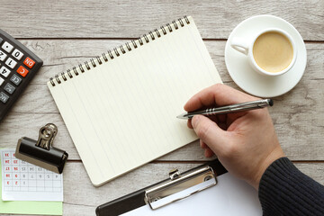 Blank notepad, coffee on brown wooden table at office workplace. male hand writes text. Top view with copy space for your design.