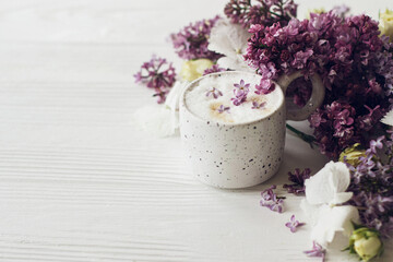 Fototapeta na wymiar Delicious coffee with lilac petals, rose and lilac branch on rustic white wooden background. Hello spring. Space for text. Happy mothers day. Good morning still life