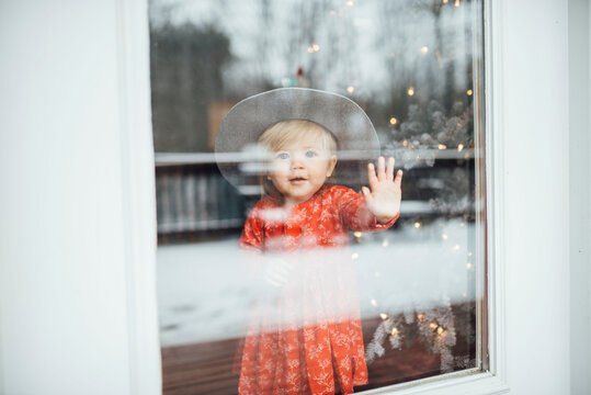 Baby girl standing and looking out window with hat by Christmas tree