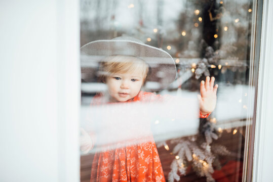 Baby girl standing and looking out window with hat by Christmas tree
