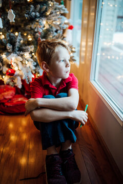 Young boy smiling and sitting by Christmas tree, looking out window