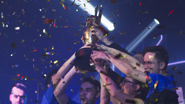 Excited gamer woman professional esportsman winner raising golden cup over head while celebrating victory in gaming tournament with male teammates