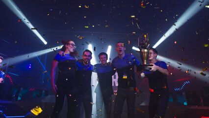 Fototapeta na wymiar Professional cybersport team of gamers winners with golden trophy hugging and jumping amidst confetti and pyrotechnics while celebrating victory in esports competition