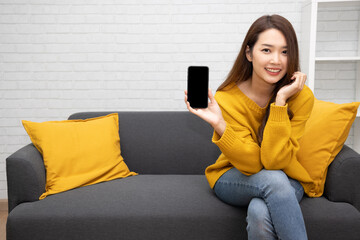 Young elegant beautiful Asian woman smiling and showing screen on mobile phone and sitting on sofa...