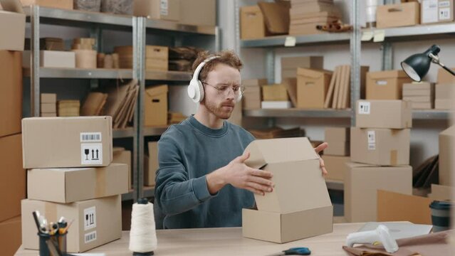 Attentive caucasian man wearing glasses and headphones sitting at the table and packing parcels at the cupboard boxes at his home office. Small business concept.