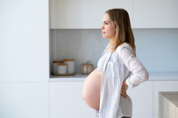 A pregnant woman stands at home in her kitchen and looks out the window.