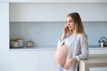 Happy pregnant woman is sitting at home in the kitchen with a mobile phone in her hands.