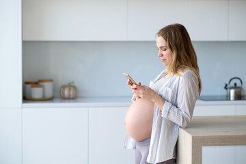 Happy pregnant woman is sitting at home in the kitchen with a mobile phone in her hands.