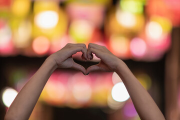 girl makes heart-shaped hand sign on blurred background of lights to symbolize friendship and...