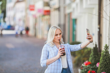 Stylish blonde woman in casual light clothes with a cocktail in her hands makes a selfie. Portrait of a beautiful fashionable woman on the street of a European city. Warm time. Shopping.