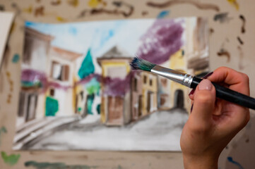 Brush in hand above watercolor painting. Colorful medieval city street on paper board.