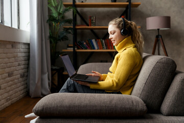 Obraz premium Young girl student studying at living room, sitting on sofa and using headphones and laptop.