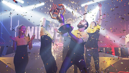 Bearded leader of cybersport team kissing golden trophy and dancing with team under confetti while...
