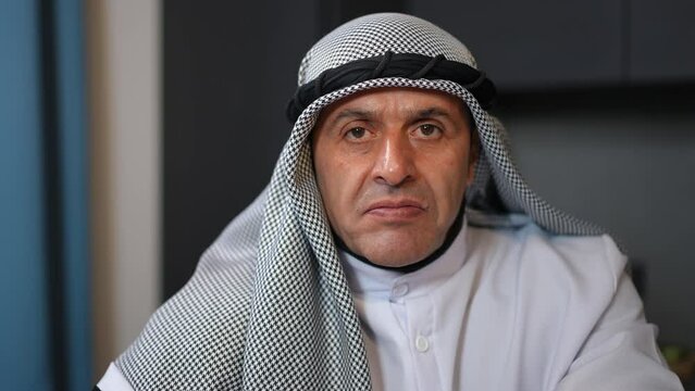 Close-up portrait of angry Middle Eastern man looking at camera with dissatisfied facial expression. Troubled irritated annoyed guy in traditional clothing posing indoors in kitchen at home