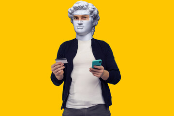 Abstract modern collage. The man with the plaster head of David Cheerful sports red-haired guy conducts online payment with smartphone on a yellow background.
