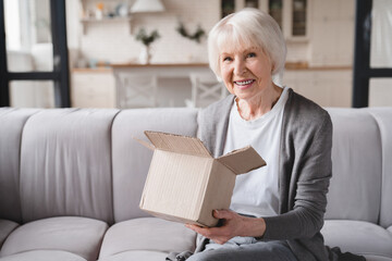 Happy smiling old elderly senior woman grandmother opening receiving post parcel delivery, presents gifts, ordering goods online on Internet looking at camera at home. - obrazy, fototapety, plakaty