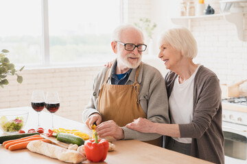 Happy caucasian active seniors grandparents loving elderly couple spouses helping cooking romantic dinner, drinking wine, making vegetable salad together at home kitchen, celebrating anniversary