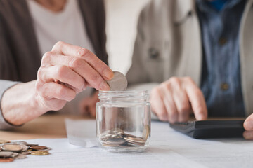 Focused closeup cropped photo of senior couple family spouses counting on calculator, savings for nest egg, putting coin into money box, economy for paying domestic bills, mortgage loan at home.