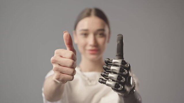 Slowmo closeup portrait of young beautiful Caucasian woman with bionic arm showing thumbs up standing on gray background