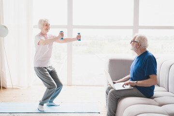 Fototapeta premium Old elderly senior grandparents training together, husband working using laptop while his wife stretching, doing physical exercises with dumbbells at home on lockdown
