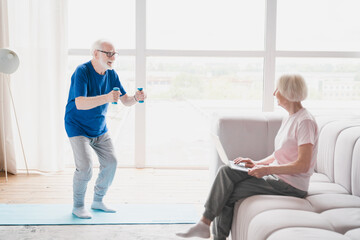 Fototapeta premium Old elderly senior grandparents training together, wife working using laptop while her husband stretching, doing physical exercises with dumbbells at home on lockdown