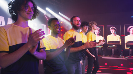Multiracial young men in yellow team t shirts waving and clapping hands while taking part in modern gaming tournament - obrazy, fototapety, plakaty