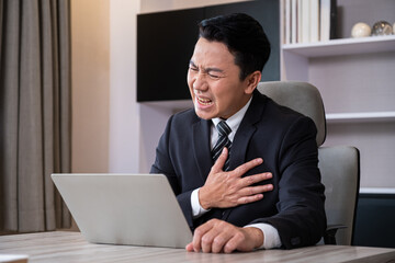 Adult men business person feeling painful from chest or heart attack while working, online meeting in front of the computer inside of the office area. Office syndrome, over working illness.