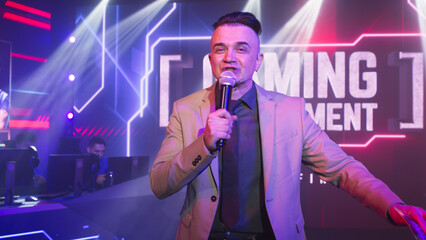 Host with microphone gesticulating and looking at camera while standing between tables with teams...