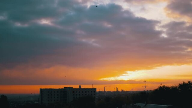 Time Lapse Sunrise over an Orange-Red Horizon and Gray Solid Clouds. Clouds slow move over treetops and unfinished buildings against the backdrop of working factories. Smoke over industrial town. 4K.
