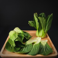 Organic green Baby Bok Choy or Brassica rapa chinensis on wooden plate on black background. Popular Chinese vegetable for Asian cuisine.Healthy vegetarian and vegan eating. - obrazy, fototapety, plakaty