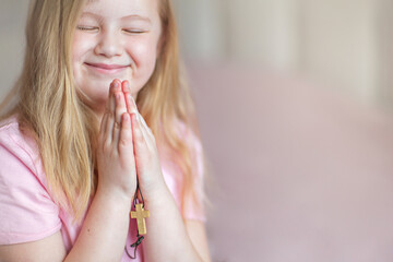 A blonde little girl, smiling, praying at home in the morning with folded hands and holding a cross.  Children's prayer, faith and prayer to God.