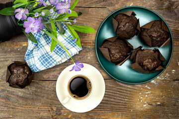 homemade chocolate muffins or madeleine in green plate with cup of coffee and pot of purple flowers on wooden table