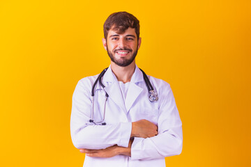 Portrait of smiling young male doctor with stethoscope around neck standing with arms crossed in white coat isolated on yellow background