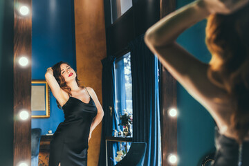 Sexy young woman in elegant cocktail silk black dress dancing and posing in mirror reflection in luxury interior. Fashionable and self-confident lady. Celebrity, superstar lifestyle. Selective focus