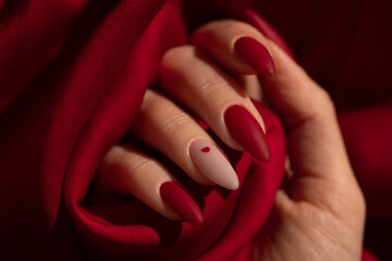 Matte red nails with small red heart on beige colour nail on the red fabric background. Saint...