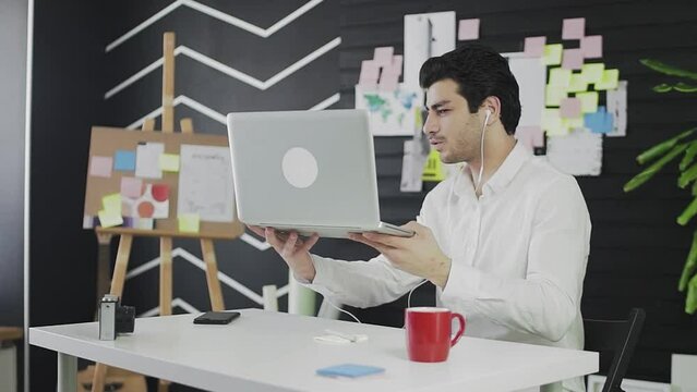 A young man of Caucasian appearance is sitting at a computer and working remotely. A young man wearing headphones communicates via video link and shows his notes on the wall