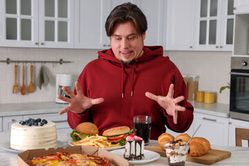 Hungry overweight man at table with sweets and fast food in kitchen