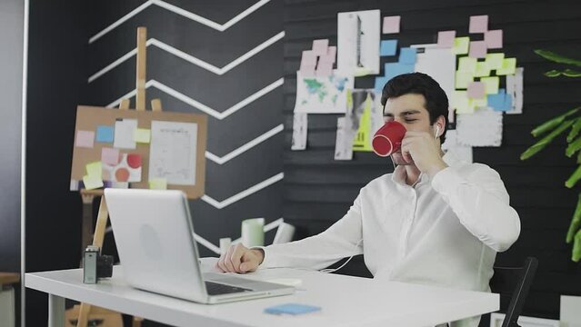 A young man of Caucasian appearance is sitting at a computer and working remotely. A young man wearing headphones communicates via video link and drinks tea or coffee