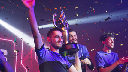 Gamer esportsman with his teammates raise and showing golden cup and waving hand near teammates clapping hands after victory in professional gaming tournament