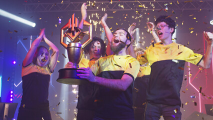 Bearded leader of cybersport team kissing golden trophy and dancing with team under confetti while celebrating victory in esports gaming championship © Framestock