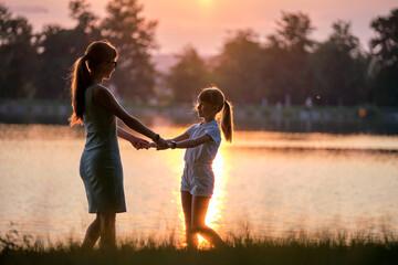 Naklejka na ściany i meble Happy mother and daughter standing together holding hands enjoying time in summer park in evening. Family love and relationship concept