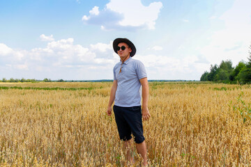 Crop and harvest. Portrait of farmer standing in gold wheat field with blue sky in background. Young man wearing sunglasses and cowboy hat in a field examining wheat crop. Oats plant