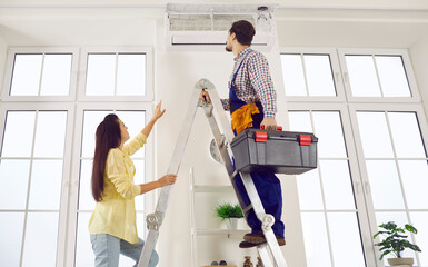 Technician repairing AC on his typical working day. Man climbs ladder with toolbox in order to check, do disinfection or fix troubles in modern white wall mounted air conditioner in young lady's home