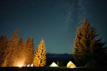 Silhouette of hikers resting besides burning bonfire near illuminated tourist tents on camping site...